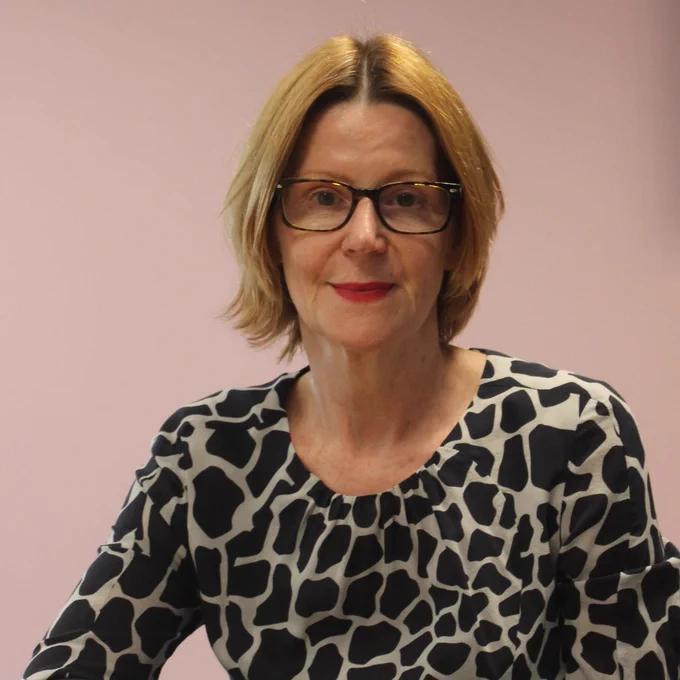 Maria has blonde hair and wears glasses and is sitting in front of a dusky pink coloured wall. Her long-sleeved top has a black and grey block pattern on it. 