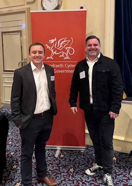 Yn y llun yma mae Guto Ifan ac Ed Gareth Poole yn sefyll ochr yn ochr o flaen baner coch Llywodraeth Cymru.  Mae'r ddau yn gwisgo crysau gwyn a'u coleri ar agor a siacedi du.  Mae Guto yn gwisgo esgidiau brown ac Ed yn gwisgo esgidiau ysgafn â carrai.