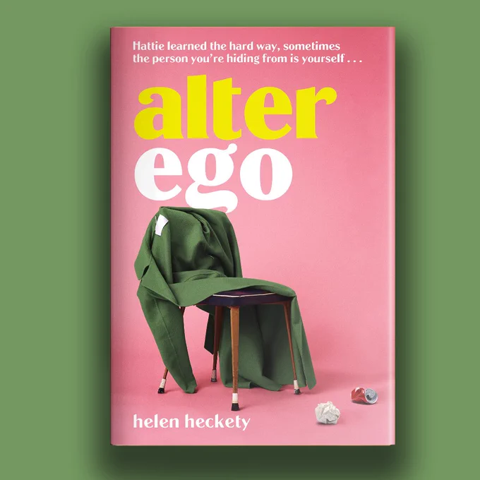 Green background with the cover of the book ALTER EGO by Helen Heckety. Cover is pink with yellow and white writing, and has a chair with a coat flung across it. Scrunched paper and tin can appear next to chair.