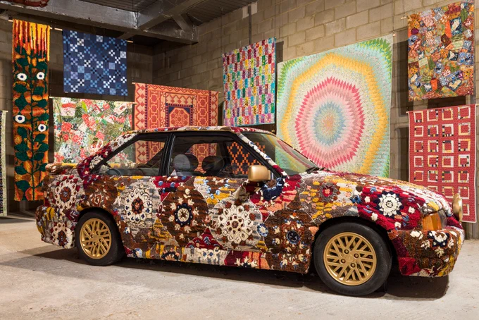A car with panels made of tufted carpet in an intricate and ornate pattern against a backdrop of colourfully patterned quilts