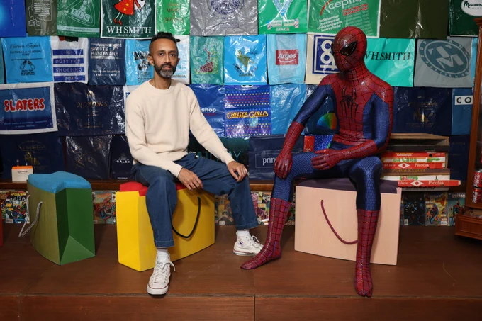 Artist Hetain Patel sitting on a stool shaped like a shopping bag, mirroring a Spiderman figure sitting next to him against a backdrop of shopping bags.