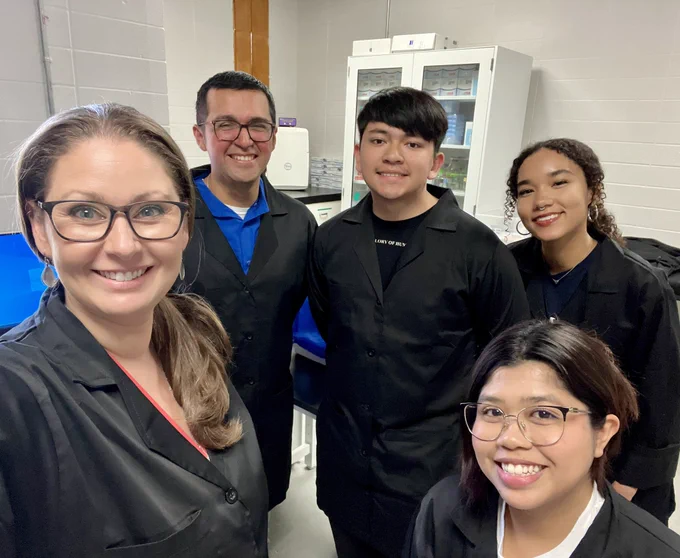 5 scientists in black lab coats smile while talking a group selfie.