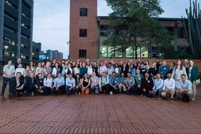 Large group of people posed for a photo on a paved area with buildings and greenery in the background at dusk.