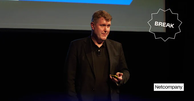 A member of the Netcompany team stands on stage looking out to an audience, in front of a dark background and projector screen.