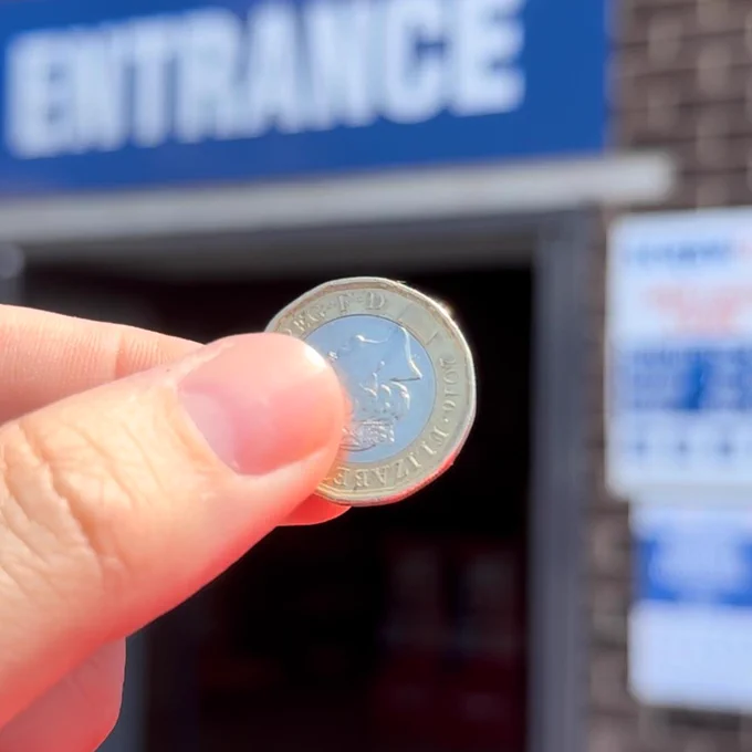 A close-up of a hand holding a £ coin in focus, with an entrance sign in the blurry background.