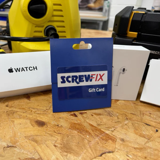 A Screwfix gift card displayed on a wooden table, surrounded by electronic devices including a vacuum and boxes labeled "Watch" and "AirPods.