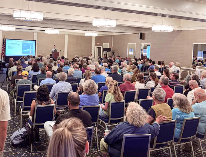 A large audience is gathered in a conference room for an information session hosted in Massachusetts about the Gulf of Mexico PSN. The room is filled with rows of seated attendees, attentively listening to a speaker at a podium in the front of the room. A presentation slide is displayed on a screen behind the speaker, detailing BOEM's Office of Renewable Energy Programs. 