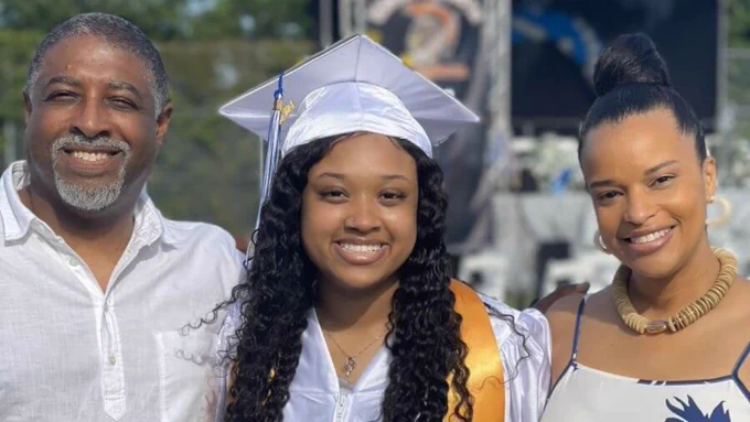 A student poses with her parents while wearing a white graduation cap and gown. 