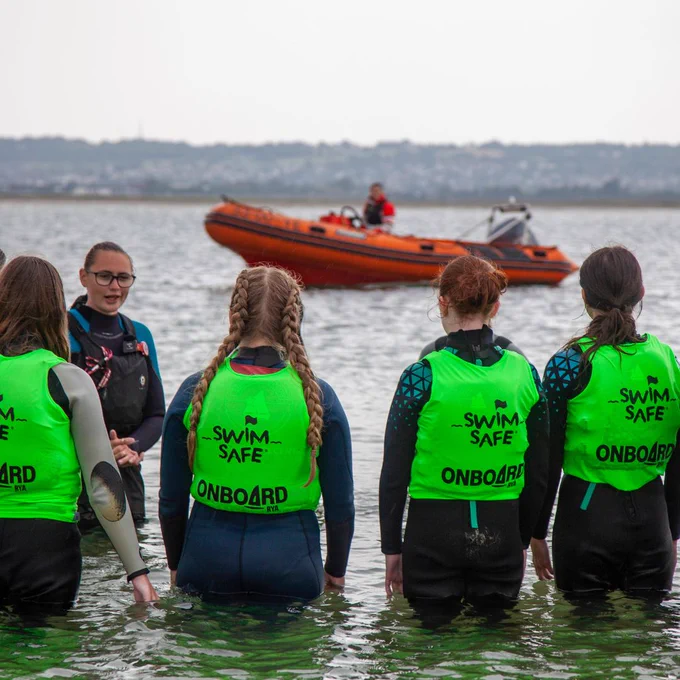 A swim safe instructor standing in water dressed in a wetsuit and life jacket, teaching a group of children wearing high visibility green vests, wetsuits and life jackets. An orange lifeboat is in the background.
