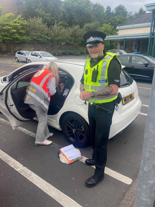 Uniform police officer standing next to a car while the Good Egg Safety Officer carries out a child car seat check, with a baby sat in its car seat.