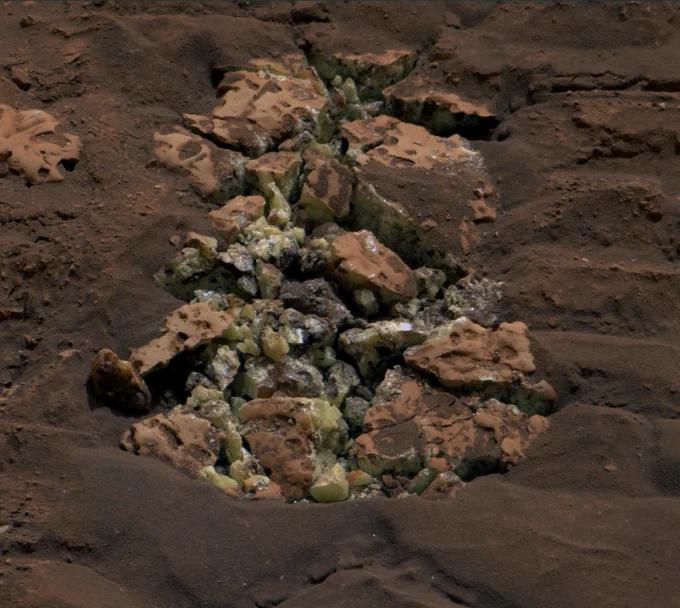 A close-up photo taken by the Curiosity rover shows pure yellow sulfur crystals within a rock the robot drove over. Surrounding the yellow crystals is sandy and rust-colored Martian rock. Along the crack in the center are dull yellow crystalline pieces of elemental sulfur.