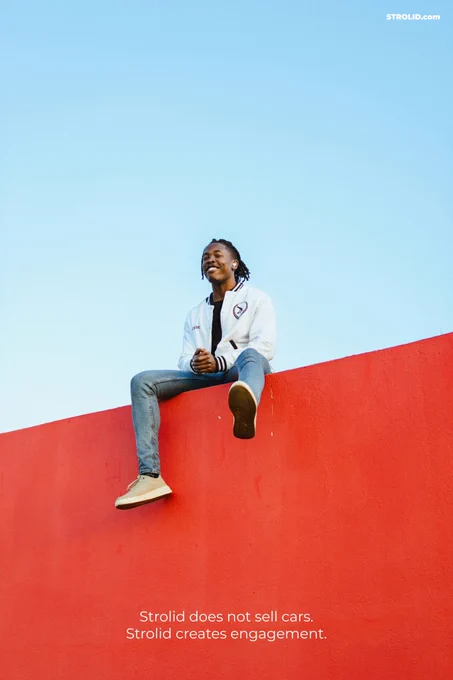 Smiling young man in a white jacket and blue jeans sitting on a bright-red wall under a blue sky.