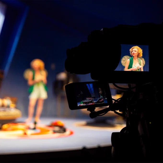 A camera focuses on a woman stood on stage. She wears a fairy dress and wings.