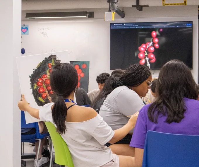 In a classroom scene in the Center for Creativity, a person holds up a large painting of Mokha beans as she compares it to the original photo of the beans on a large TV screen. Other students continue working on their own paintings.