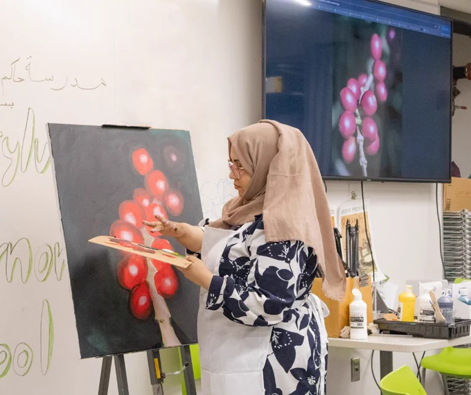 Haifa Subay, wearing a hijab and a white apron, paints Mokha beans in front of the classroom next to a digital display of Mokha beans that the class is painting together.