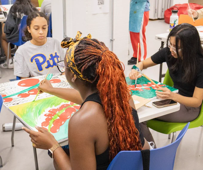Three students, including one with a Pitt T-shirt, work on their paintings of the Mokha beans in a variety of different styles. Art supplies are scattered around.