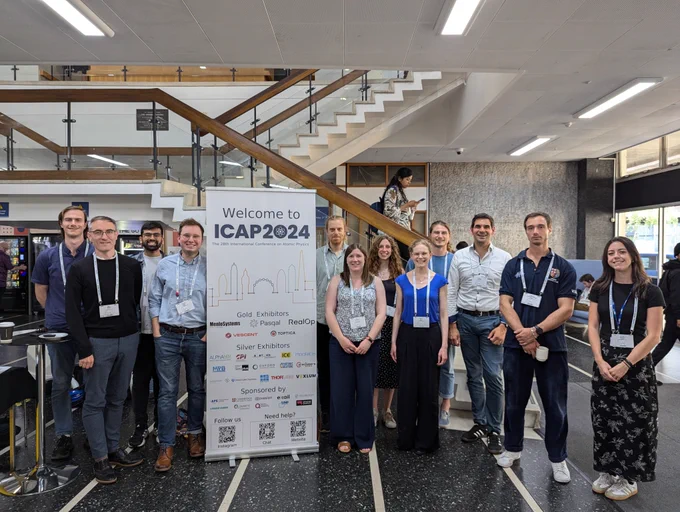 A group of people stand around a sign reading 'Welcome to ICAP 2024' in the entrance foyer of a university building with staircases crossing behind them. Most of them look happy to be there!