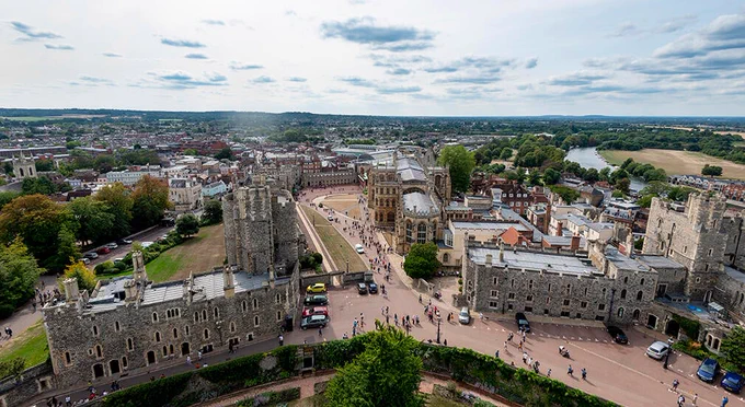Aerial view of Windsor Castle with surrounding grounds and a suburban area in the background under a cloudy sky.