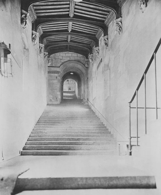 Black and white photograph of a stone staircase inside a historical building, featuring high ceilings with exposed wooden beams, leading upward to a softly lit archway at the top.