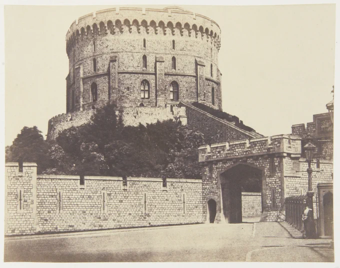 A historical photograph of Windsor Castle's Round Tower viewed from an adjacent gateway. The image shows the textured stone facade of the large circular tower and a part of the stone gateway, revealing a clear sky in the background.