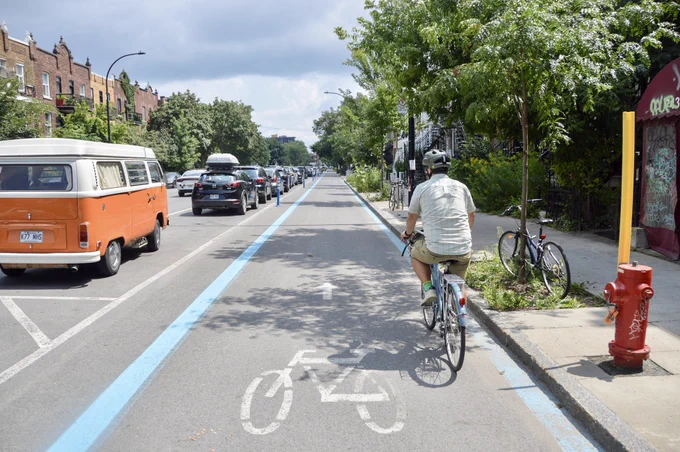 A man in casual summer attire rides a bicycle along a wide protected bike lane along a commercial shopping street in Montreal.