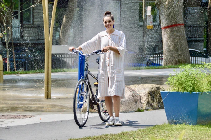 A young woman in a white dress poses for a portrait with her bicycle in a spray park in the north of Montreal.
