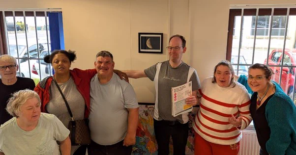 Image of group of adults, all smiling, from left: man wearing black top, woman wearing grey top, woman wearing grey top and red jacket, man wearing grey top, man wearing grey and white top and glasses, woman wearing white and red striped top, woman wearing black top and green cardigan.