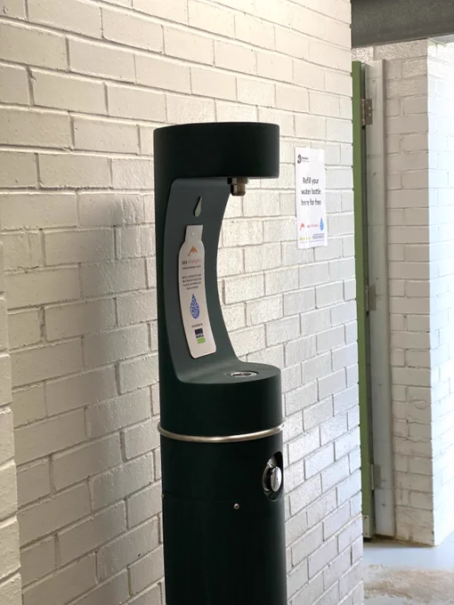 Side view of a black cylindrical water station with an inlet to stand your water bottle in and fill it. It is stood against a white brick wall. (c) Clare Galpin