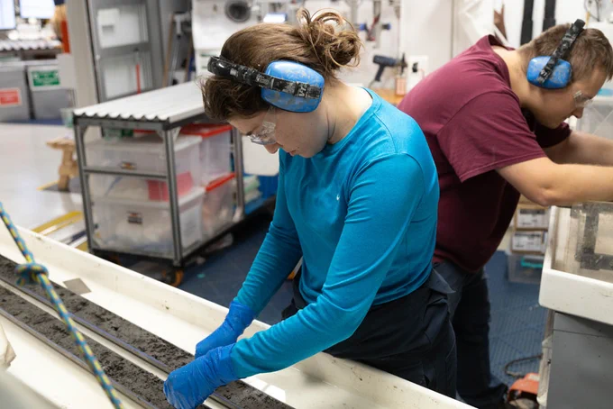 Brittany Stockmaster (Marine Laboratory Specialist, IODP JRSO), front, cleans the surface of a freshly split core while Max Witek (Marine Laboratory Specialist, IODP JRSO), back, cuts a dropstone in half. (Credit: Daniel Rudbäck, IODP JRSO) 