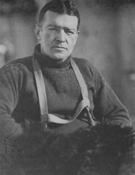 Black and white portrait of Ernest Shackleton. Looking directly at camera, Shackleton is seen seated, wearing a thick woollen jumper with reindeer fur gloves draped around his neck.