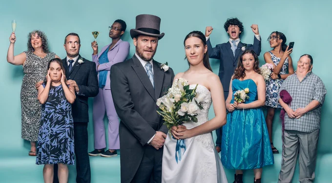 Against a pale blue background, a group of wedding guests in formal clothing are gathered, some raising a glass. In the centre, a bride and groom stand together and look slightly apprehensive. 