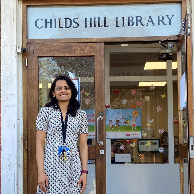 Library manager Neha Dhakar, wearing a white patterned dress, stands in front of an open door above which is a sign reading 'Childs Hill Library'
