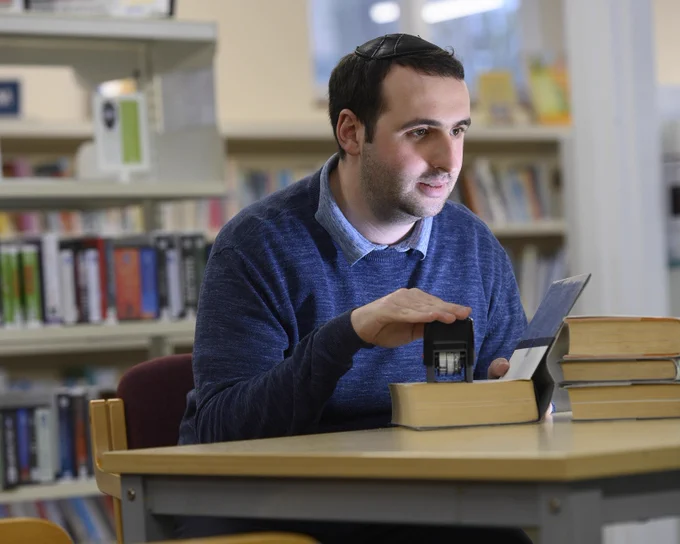 Library volunteer Shammai Rotenberg , wearing a dark blue jumper, sits at a table stamping books in Childs Hill Library