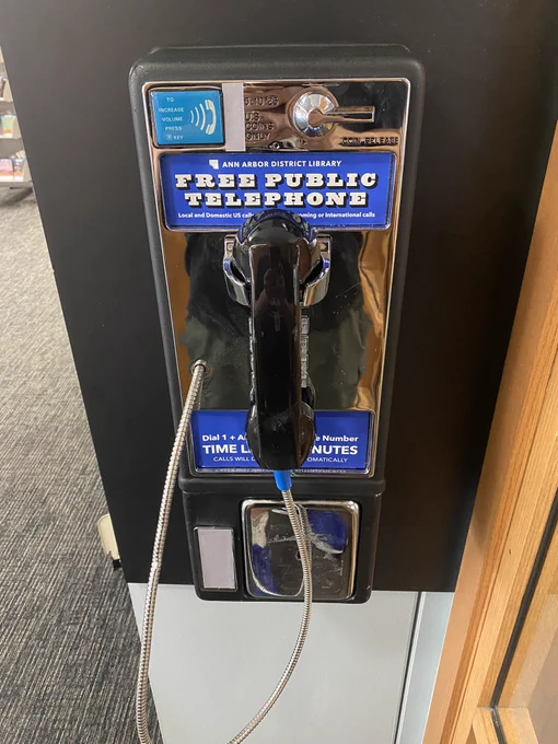 A classic pay phone mounted on the wall of the downtown library by the hold shelves. The coin slot is taped over, and a sticker reads “Ann Arbor District Library Free Public Telephone”