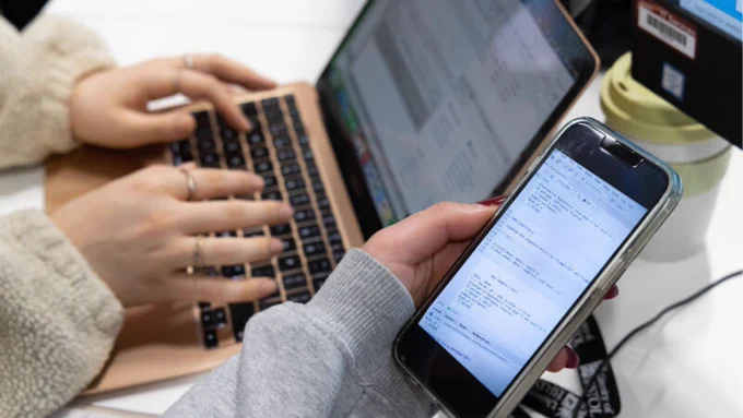 Student holding a phone next to a laptop screen with another student typing at that laptop