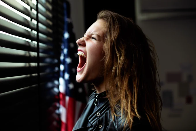 Election anxiety: Young woman in front of a window with the blinds drawn, next to an American flag, screaming with frustration over uncertainty and political stress.