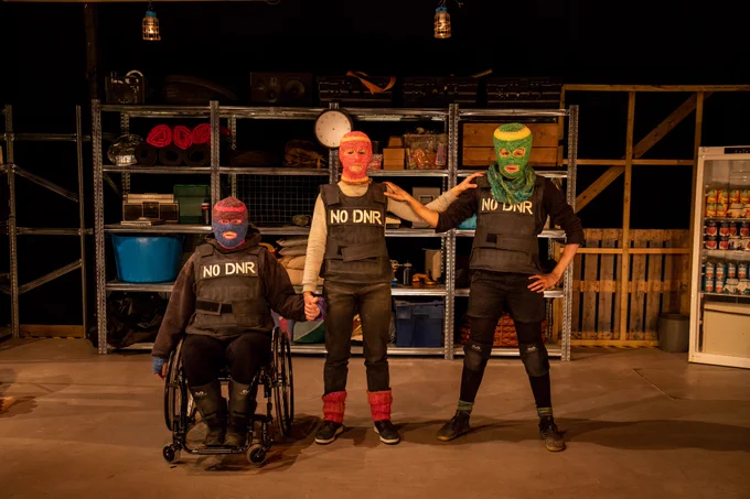 Three people stare directly at the camera and are each wearing different coloured, knitted balaclavas where only their eyes and mouths are exposed. They are all wearing black stab vests with the words ‘NO DNR’ stencilled onto the vests in white paint. They are holding hands. Jess is on the left of the image. She is sitting in a wheelchair and wearing a blue and pink knitted balaclava. Charmaine is on the right of the image. She is wearing a knitted green balaclava and blue knitted snood. She has a hand on her hip. Chopin is in the centre of the image between Jess and Charmaine. She is wearing a red and orange knitted balaclava. In the background is a shelving unit with miscellaneous items including old computers, storage boxes and rolled blankets. 