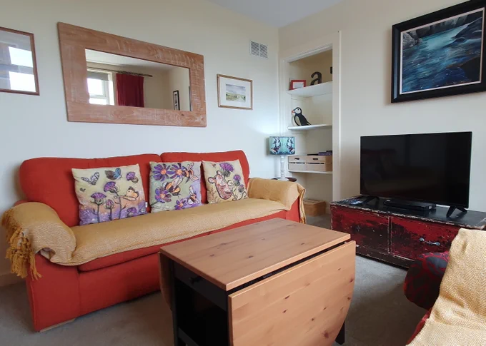 A living room with a three seater sofa, a coffee table, a TV, shelves and a mirror