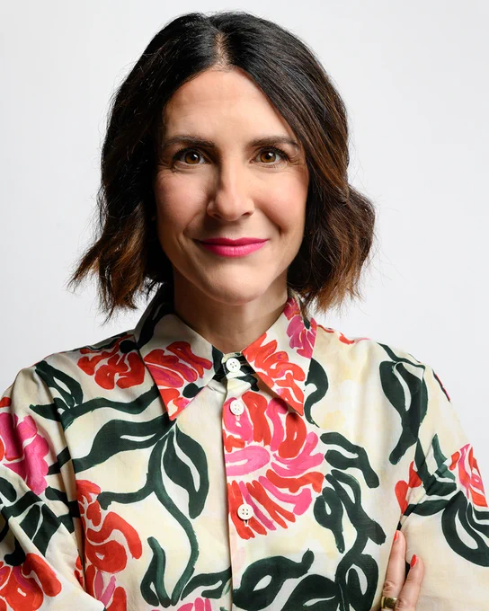 A portrait of a white woman, Kelly Shindler, who is wearing a cream colored blouse with a floral pattern of red and pink flowers and dark green foliage. Her brown hair sits a bit higher than her shoulders and is parted at the center.
