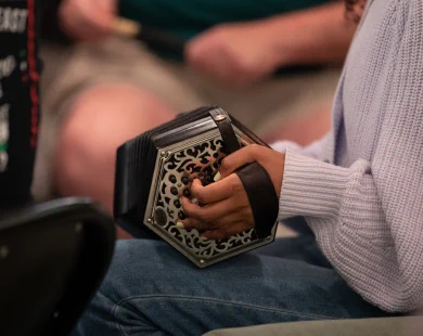 A person in a light purple sweater and jeans plays the concertina.