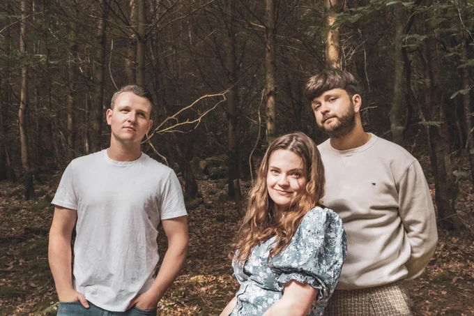 Three people in light colored casual clothing stand together and pose for the camera in the woods.