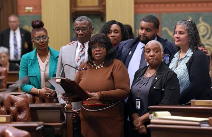 Michigan state Representative Brenda Carter speaking on House floor. Reps. Helena Scott, Felicia Brabec, Donovan McKinney, Stephanie A. Young, Amos O'Neal and Cynthia Neeley are standing behind her in support.