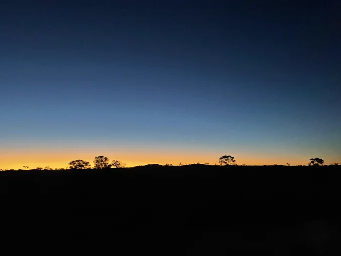 Photo of a lab almost flat horizon with a couple of small gum trees. The horizon is black, with an orange sunset that fades into a deep blue night sky.