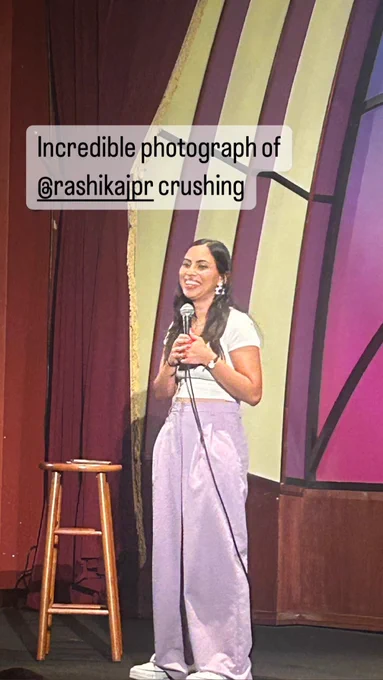 Rashika Jaipuriar stands on stage talking into a microphone.