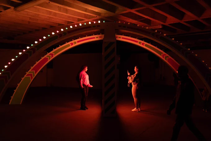 Two individuals interact with an illuminated, colorful archway installation in a dimly lit room. One person stands under the arch while another appears to take a photo.