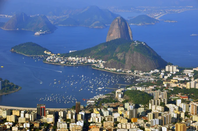 Carioca landscape of Rio de Janeiro. A tall peak of granite is sugar loaf mountain. surrounded by other hills and a broad bay of water. lots of buildings are in the foreground. 