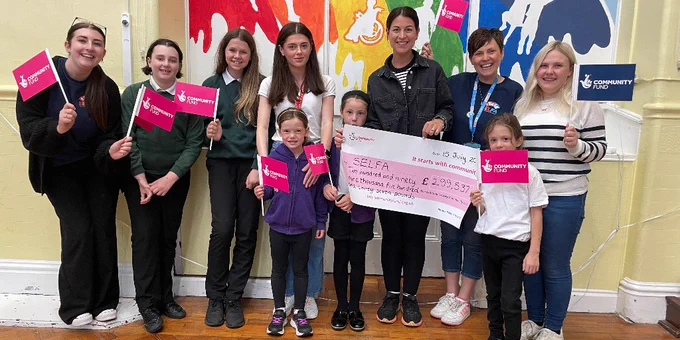 Children and young people holding giant National Lottery cheque and waving National Lottery branded flags.