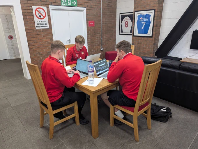 3 Apprentices sat around a table, all wearing red, working as a group on laptops to complete their apprenticeship work. 