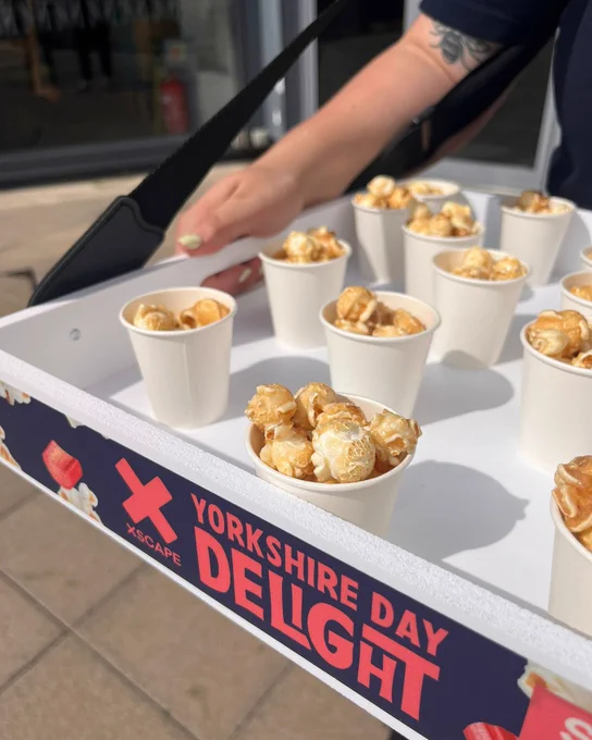 A person serves sample cups of popcorn from a tray labeled "Yorkshire Day Delight" at an outdoor event.