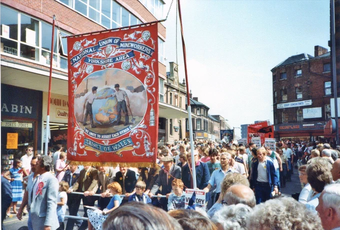 Image is of the Miners' Gala Parade that took place in Wakefield on 16 June 1984. The Prince of Wales Branch Yorkshire Area banner can clearly be seen which carries the slogan 'WE UNITE TO ASSIST EACH OTHER'.